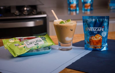 Copo com bebida cremosa e limão ao lado de um sachê azul de Nescafé Ice e embalagem verde sobre tecido azul.