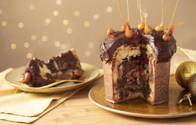 Fotografia em tons de amarelo em uma bancada de madeira clara, um pano amarelo e decoração natalina de fundo. Ao centro, um prato dourado com o panettone de avelã recheado sem um pedaço. Ao lado, um prato com a fatia do panettone. Fotografia em tons de amarelo em uma bancada de madeira clara, um pano amarelo e decoração natalina de fundo. Ao centro, um prato dourado com o panettone de avelã recheado sem um pedaço. Ao lado, um prato com a fatia do panettone.