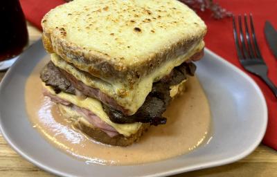 Foto da receita de Francesinha. Oberva0se um prato de cerâmica cinza com o sanduíche por cima, com queijo gratinado e um molho amarronzado.