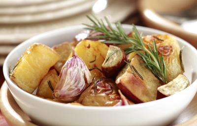 Fotografia em tons de bege em uma bancada de madeira escura, uma toalha vinho, um pote bege e um branco redondo em cima com o mix de batatas assadas dentro dele. Fotografia em tons de bege em uma bancada de madeira escura, uma toalha vinho, um pote bege e um branco redondo em cima com o mix de batatas assadas dentro dele.