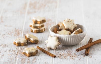 Fotografia de várias unidades de biscoito alemão em formato de estrela e uma pasta branca por cima. Uma parte dos biscoitos estão dentro de um pote de vidro branco, e o resto está espalhado em volta do refratário, sobre uma mesa de madeira branca.