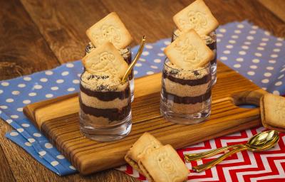 Imagem de uma bancada de madeira na qual há dois panos em tons azul claro e vermelho com texturas de bolinhas e listras. Há uma tábua pequena e sobre ela quatro copinhos com a receita em camadas de creme e biscoito triturado.