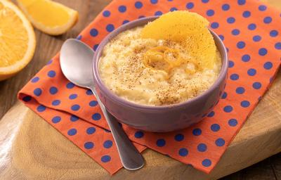 Foto em tons de laranja da receita de arroz-doce de laranja servida em um bowl de porcelana lilás sobre um pano laranja com bolinhas azuis com uma colher prateada ao lado. Ao fundo há laranjas cortadas sobre uma tábua de madeira Foto em tons de laranja da receita de arroz-doce de laranja servida em um bowl de porcelana lilás sobre um pano laranja com bolinhas azuis com uma colher prateada ao lado. Ao fundo há laranjas cortadas sobre uma tábua de madeira