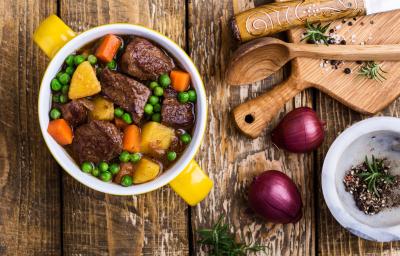 Fotografia vista de cima de um cozido de carne com ervilhas frescas, legumes e caldo de carne. Ao lado, duas cebolas roxas, uma tábua e colher de madeira e alguns temperos que estão dentro de um refratário fundo e pequeno, sobre uma mesa de madeira. Fotografia vista de cima de um cozido de carne com ervilhas frescas, legumes e caldo de carne. Ao lado, duas cebolas roxas, uma tábua e colher de madeira e alguns temperos que estão dentro de um refratário fundo e pequeno, sobre uma mesa de madeira.