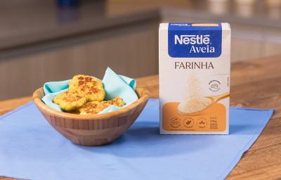Fotografia em tons claros de uma mesa de madeira com um pano azul. Sobre o pano tem um recipiente de madeira fundo com um paninho azul claro com alguns nuggets saudáveis, ao lado da embalagem de NESTLÉ Farinha de Aveia.
