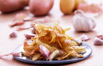 Foto da receita de casca de batata no alho, servida em um prato azul, decoradas com dentes de alho, numa mesa clara com mais batatas e alhos inteiros ao fundo
