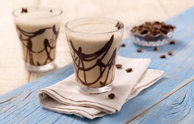 Foto da receita de smoothie de cappuccino e coco. Observa-se dois copos transparentes com calda de chocolate e a bebida, sobre um guardanapo bege em uma superfície de madeira metade bege e metade azul clara. Como decoração, grãos de café no fundo