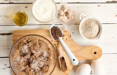 Fotografia em tons de branco e marrom de uma bancada branca com uma tábua de madeira clara, sobre ela um recipiente de vidro com ingredientes. Ao lado dois ovos, e recipientes com azeite, iogurte, fermento e aveia.