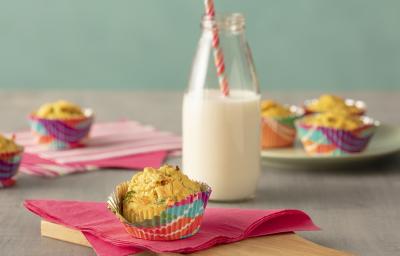 Fotografia em tons de rosa, em uma bancada de madeira de cor cinza. Ao centro, uma tábua de madeira com um pano rosa em cima. Em cima do pano está o muffin e ao lado há um pote de vidro contendo leite. AO fundo alguns muffins espalhados. Fotografia em tons de rosa, em uma bancada de madeira de cor cinza. Ao centro, uma tábua de madeira com um pano rosa em cima. Em cima do pano está o muffin e ao lado há um pote de vidro contendo leite. AO fundo alguns muffins espalhados.
