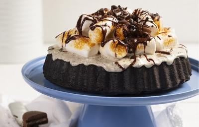 Fotografia em tons de branco, em uma bancada branca de madeira. Ao centro, um boleiro azul contendo a torta s'more. Ao lado, uma faca e ao fundo um vaso branco de flores amarelas. Fotografia em tons de branco, em uma bancada branca de madeira. Ao centro, um boleiro azul contendo a torta s'more. Ao lado, uma faca e ao fundo um vaso branco de flores amarelas.