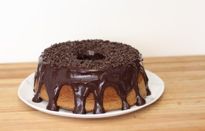 Bolo de chocolate com furo no meio, em um prato branco. O bolo é coberto com uma camada de cobertura de chocolate brilhante que escorre pelas laterais. O topo é enfeitado com granulado de chocolate. O bolo está sobre uma superfície de madeira. Bolo de chocolate com furo no meio, em um prato branco. O bolo é coberto com uma camada de cobertura de chocolate brilhante que escorre pelas laterais. O topo é enfeitado com granulado de chocolate. O bolo está sobre uma superfície de madeira.