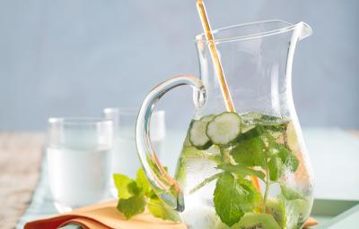 Fotografia em tons de verde e cinza, com jarra de vidro ao centro com água, kiwi, pepino e hortelã sobre bandeja em tom de verde com guardanapo laranja, ao lado dois copos com água, tudo sobre toalha em tons de azul. Fotografia em tons de verde e cinza, com jarra de vidro ao centro com água, kiwi, pepino e hortelã sobre bandeja em tom de verde com guardanapo laranja, ao lado dois copos com água, tudo sobre toalha em tons de azul.
