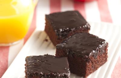 Foto com 3 pedaços quadrados de bolo de Nescau e cobertura de Nescau. No fundo da foto, há uma jarra com suco de laranja e 2 maças em um prato vermelho Foto com 3 pedaços quadrados de bolo de Nescau e cobertura de Nescau. No fundo da foto, há uma jarra com suco de laranja e 2 maças em um prato vermelho