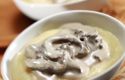 Fotografia em tons de amarelo em uma bancada de madeira, um pano bege e dois recipientes ovais brancos com a polenta coberta com molho de funghi. Fotografia em tons de amarelo em uma bancada de madeira, um pano bege e dois recipientes ovais brancos com a polenta coberta com molho de funghi.