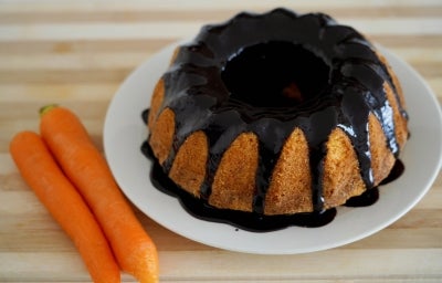 Bolo de cenoura inteiro, assado em forma de buraco no meio, está sobre um prato branco. Ele está coberto com uma generosa calda de chocolate escorrida pelas laterais. Ao lado do prato, há duas cenouras cruas, inteiras.. Bolo de cenoura inteiro, assado em forma de buraco no meio, está sobre um prato branco. Ele está coberto com uma generosa calda de chocolate escorrida pelas laterais. Ao lado do prato, há duas cenouras cruas, inteiras..