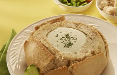 Fotografia em tons de amarelo, branco e bege, com prato branco contendo pão italiano recheado com creme, dois espetos com legumes e um guardanapo verde ao lado, ao fundo potinhos com legumes, espetinhos e pão, tudo sobre toalha amarela.