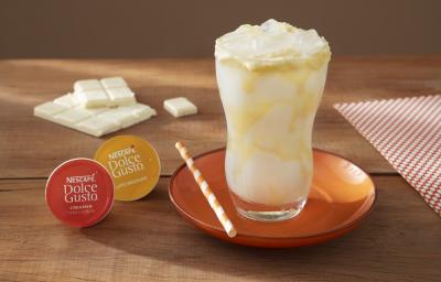 Foto da receita de mocha branco com latte macchiato servida em um copo de vidro alto sobre um pratinha laranja em cima de uma mesa de madeira com uma barra de galak e um paninho ao fundo