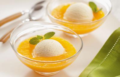 Fotografia em tons de amarelo e verde em uma bancada de madeira clara, um pano verde, um prato redondo branco com duas taças de vidro com o creme de manga e bolas de sorvete de creme em cada uma delas.