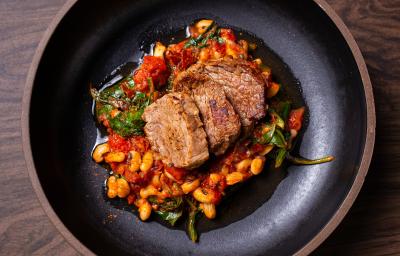 Fotografia de um prato grosso fundo preto com três fatias grossas de carne de sol sobre um molho de tomate e por baixo tem feijão manteiguinha e espinafre. O prato está sobre uma bancada de madeira.