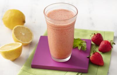 Fotografia em tons de branco, roxo e verde de uma bancada de madeira branca e um paninho verde, sore ele um apoio de copo quadrado roxo com um copo de vidro com o suco de morango. Ao lado três morangos e dois limões. Fotografia em tons de branco, roxo e verde de uma bancada de madeira branca e um paninho verde, sore ele um apoio de copo quadrado roxo com um copo de vidro com o suco de morango. Ao lado três morangos e dois limões.