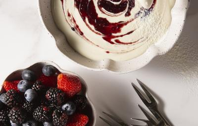 Fotografia em tons de branco, em uma mesa de madeira de cor branca. Ao centro, um recipiente contendo o fondue e ao lado um outro recipiente contendo as frutas. Ao fundo, alguns garfos. Fotografia em tons de branco, em uma mesa de madeira de cor branca. Ao centro, um recipiente contendo o fondue e ao lado um outro recipiente contendo as frutas. Ao fundo, alguns garfos.