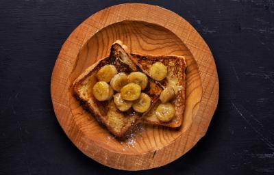 Fotografia em tons de madeira com um prato de madeira ao centro. Em cima do prato existe dois pães de forma cobertos com banana caramelizada e mel