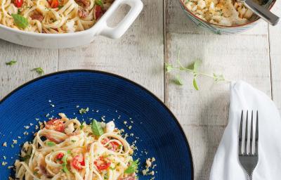 Fotografia em tons de azul em uma mesa de madeira clara com um guardanapo branco, um prato azul redondo com o macarrão com tomates e anchova.