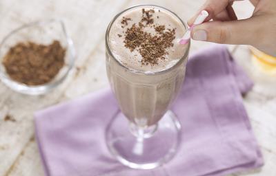 Fotografia em tons e branco e roxo de uma bancada branca com um paninho roxo, sobre ele uma taça de vidro com milk shake e um canudo. Ao lado um recipiente de vidro com raspas de chocolate.