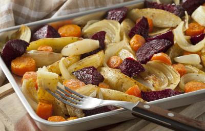 Fotografia em tons de amarelo e roxo, embaixo um pano quadriculado branco e bege, sobre ele uma assadeira de inox com batatas, cenouras, cebolas e beterrabas, há um garfo com cabo de madeira espetando uma batata. Fotografia em tons de amarelo e roxo, embaixo um pano quadriculado branco e bege, sobre ele uma assadeira de inox com batatas, cenouras, cebolas e beterrabas, há um garfo com cabo de madeira espetando uma batata.