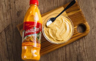 Fotografia em tons de amarelo em uma bancada de madeira, um potinho de vidro com uma mousse de maracujá com uma colher dentro e uma embalagem de um suco de maracujá Maguary ao lado. Fotografia em tons de amarelo em uma bancada de madeira, um potinho de vidro com uma mousse de maracujá com uma colher dentro e uma embalagem de um suco de maracujá Maguary ao lado.