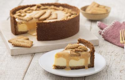 Foto da receita de Cheesecake de Tostines Especiarias. Observa-se uma torta com um pedaço tirado em um prato de sobremesa branco. A calda de doce de leite está escorrendo na fatia e biscoitos TOSTINES estão no topo decorando.