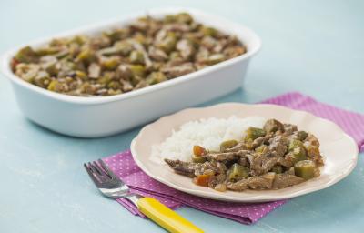 Fotografia em tons de azul e rosa em uma bancada de madeira azul, um guardanapo de pano rosa com bolinhas brancas, um refratário branco retangular com o picadinho de carne com quiabo. Ao lado, um prato rosa redondo com uma porção do picadinho e arroz. Fotografia em tons de azul e rosa em uma bancada de madeira azul, um guardanapo de pano rosa com bolinhas brancas, um refratário branco retangular com o picadinho de carne com quiabo. Ao lado, um prato rosa redondo com uma porção do picadinho e arroz.