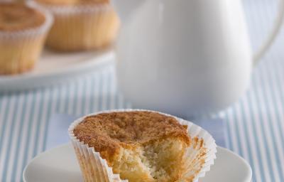 Fotografia em tons de azul em uma mesa de madeira com uma toalha listrada azul, um paninho azul, um pratinho de sobremesa branco com o muffin de banana com iogurte. Ao fundo, um bule branco com chá dentro. Fotografia em tons de azul em uma mesa de madeira com uma toalha listrada azul, um paninho azul, um pratinho de sobremesa branco com o muffin de banana com iogurte. Ao fundo, um bule branco com chá dentro.