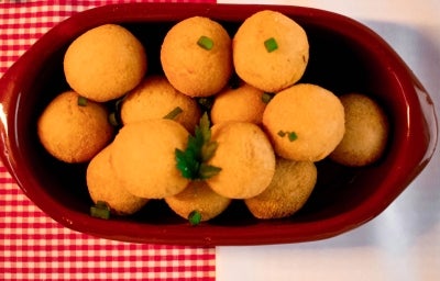 Fotografia vista de cima de bolinhas de queijo dentor de um recipiente oval marrom de vidro sobre um pano vermelho quadriculado com branco, o qual está apoaido em uma mesa branca. Fotografia vista de cima de bolinhas de queijo dentor de um recipiente oval marrom de vidro sobre um pano vermelho quadriculado com branco, o qual está apoaido em uma mesa branca.