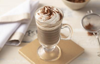 Fotografia em tons de marrom de uma bancada, ao centro uma placa quadrada bege e um copo transparente com alça e a bebida decorada com chantilly e chocolate em pó.