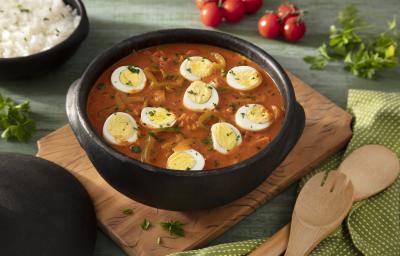 Fotografia em tons de verde em uma bancada de madeira verde, uma tábua de madeira, um paninho verde com bolinhas brancas, uma panela de barro grande ao centro com a moqueca de ovos dentro dela. Ao fundo, um recipiente com arroz, tomates e salada verde. Fotografia em tons de verde em uma bancada de madeira verde, uma tábua de madeira, um paninho verde com bolinhas brancas, uma panela de barro grande ao centro com a moqueca de ovos dentro dela. Ao fundo, um recipiente com arroz, tomates e salada verde.