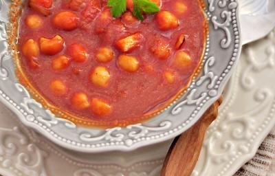 Fotografia vista de cima de uma sopa de tomates com nhoquettes em um prato fundo desenhado em tons de cinza, apoiado sobre um prato raso branco desenhado com uma colher apoiada. Os pratos estão sobre uma toalha de mesa marrom. Fotografia vista de cima de uma sopa de tomates com nhoquettes em um prato fundo desenhado em tons de cinza, apoiado sobre um prato raso branco desenhado com uma colher apoiada. Os pratos estão sobre uma toalha de mesa marrom.