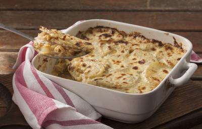 Fotografia em tons de marrom, branco e rosa de uma bancada marrom vista de frente. Contém um recipiente quadrado branco com alças e batatas e queijo gratinado, por cima uma colher com um pedaço da receita e ao lado um pano listrado nas cores rosa e branco Fotografia em tons de marrom, branco e rosa de uma bancada marrom vista de frente. Contém um recipiente quadrado branco com alças e batatas e queijo gratinado, por cima uma colher com um pedaço da receita e ao lado um pano listrado nas cores rosa e branco