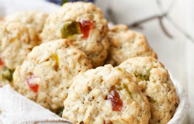 Fotografia em tons claros de cookies de massa branca e neston e frutas que estão dentro de uma cestinha de madeira forrada com um pano branco. Ao fundo, um copo de alumínio branco com desenhos em azul e verde. Tudo está sobre uma mesa de madeira clara.
