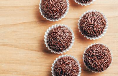 Fotografia vista de cima de seis brigadeiros passados no granulado dentro de uma forminha branca sobre uma tábua de madeira clara. Fotografia vista de cima de seis brigadeiros passados no granulado dentro de uma forminha branca sobre uma tábua de madeira clara.