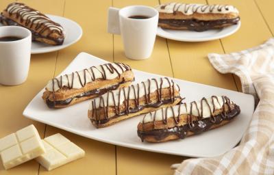 Fotografia em tons de amarelo em uma bancada de madeira com um pano xadrez brege, um recipiente retangular branco ao centro com três bombas de chocolate com cobertura de chocolate branco. Ao fundo, duas xícaras com café e dois pratinhos com duas bombas. Fotografia em tons de amarelo em uma bancada de madeira com um pano xadrez brege, um recipiente retangular branco ao centro com três bombas de chocolate com cobertura de chocolate branco. Ao fundo, duas xícaras com café e dois pratinhos com duas bombas.