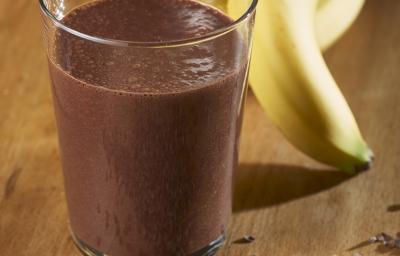 Fotografia de um copo de vidro com uma vitamina de banana e chocolate Nescau. Ao fundo, um cacho de bananas, e na frente do copo, raspas de chocolate sobre uma mesa de madeira. Fotografia de um copo de vidro com uma vitamina de banana e chocolate Nescau. Ao fundo, um cacho de bananas, e na frente do copo, raspas de chocolate sobre uma mesa de madeira.