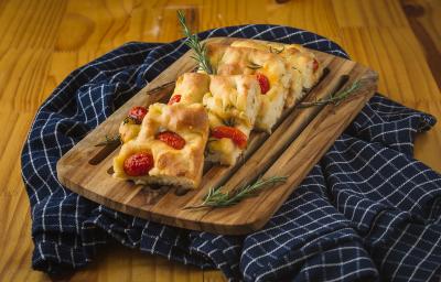 Foto aproximada de uma bancada com um tecido azul quadriculado sobre e nele uma tábua de madeira com pedaços de Focaccia, decorada com tomates-cereja e ramos de alecrim