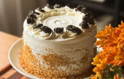Fotografia de um bolo inteiro de doce de leite com ameixa e coco, decorado com chantilly e ameixas sobre um prato grande e fundo branco.