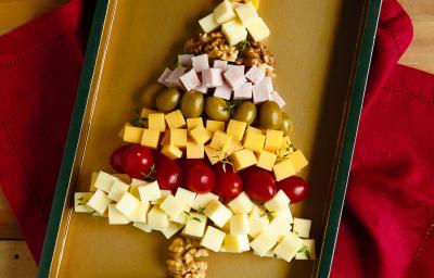 Fotografia em tons de vermelho em uma bancada de madeira escura, um pano vermelho, uma bandeja com vários frios (muçarela, presunto, queijo prato, tomatinhos e nozes) empilhados formando uma árvore de natal.