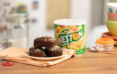 Foto de uma bancada de madeira. Sobre ela há uma tecido laranja claro e nele um prato pequeno com três brownies de cacau. Ao fundo há uma lata de Neston 3 Cereais e alguns utensílios de cozinha Foto de uma bancada de madeira. Sobre ela há uma tecido laranja claro e nele um prato pequeno com três brownies de cacau. Ao fundo há uma lata de Neston 3 Cereais e alguns utensílios de cozinha