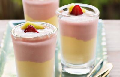 Fotografia em tons de rosa e amarelo em uma bancada de madeira, um recipiente retangular com três taças de vidro com o creme de frutas com o sorvete de creme. Morango e manga para decorar as taças.