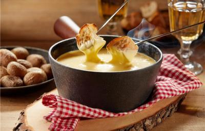 Fotografia mostra um creme brilhante com textura aveludada, servido em aparelho de fondue de cerâmica, acompanhado de cubos de pão dourados mergulhado