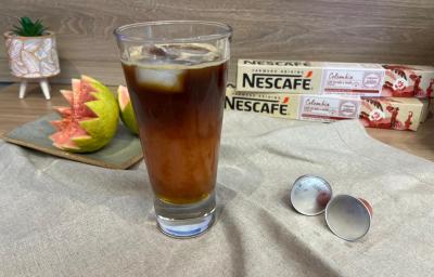 Fotografia em tons de rosa e verde com um copo alto ao centro. Dentro do copo existe uma mistura de café com suco de goiaba e cubos de gelo. Ao fundo existe uma goiaba cortada e duas caixas de cápsulas de NESCAFÉ Fotografia em tons de rosa e verde com um copo alto ao centro. Dentro do copo existe uma mistura de café com suco de goiaba e cubos de gelo. Ao fundo existe uma goiaba cortada e duas caixas de cápsulas de NESCAFÉ