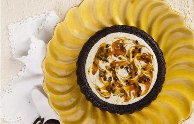 Fotografia em tons de amarelo em uma bancada de madeira branca. No centro, um prato redondo contendo o Bolo Piscina. Ao fundo, um recipiente com poupa de maracujá e um outro com negresco. Fotografia em tons de amarelo em uma bancada de madeira branca. No centro, um prato redondo contendo o Bolo Piscina. Ao fundo, um recipiente com poupa de maracujá e um outro com negresco.